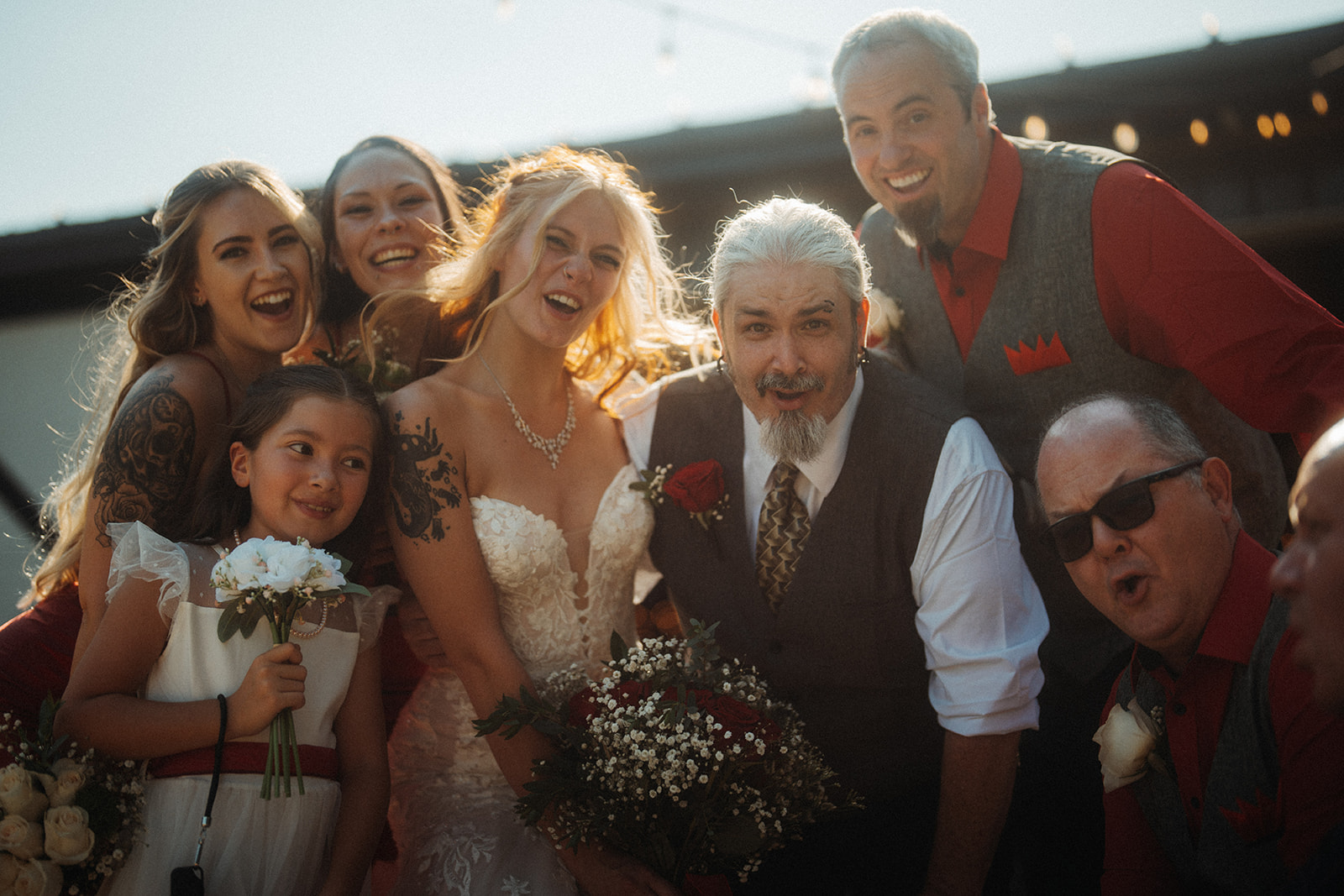 Wedding party group selfie with bride, groom, and guests smiling outdoors.